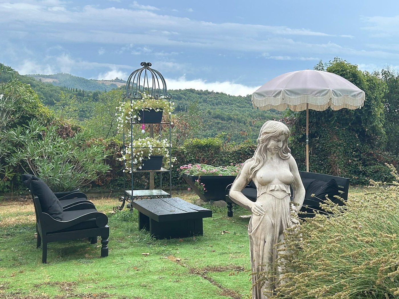 Het uitzicht vanuit de tuin met een vrouwenstandbeeld, een beige parasol, zwarte loungebanken en een zee van heuvels