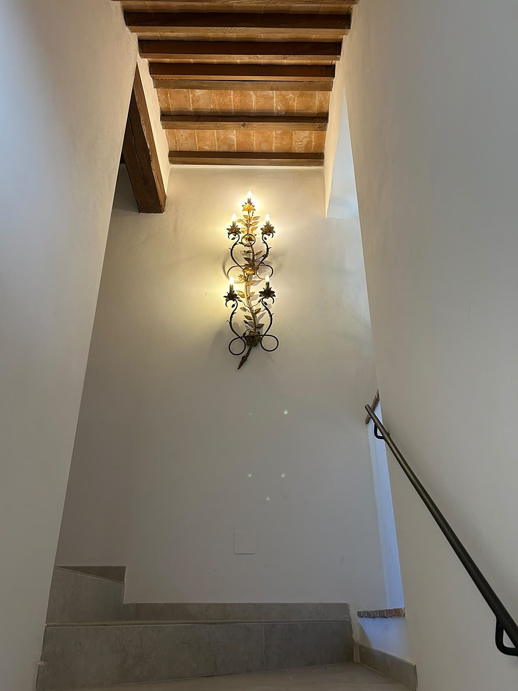 De binnentrap met witte wanden, een houten balkenplafond en een goudkleurige wandlamp