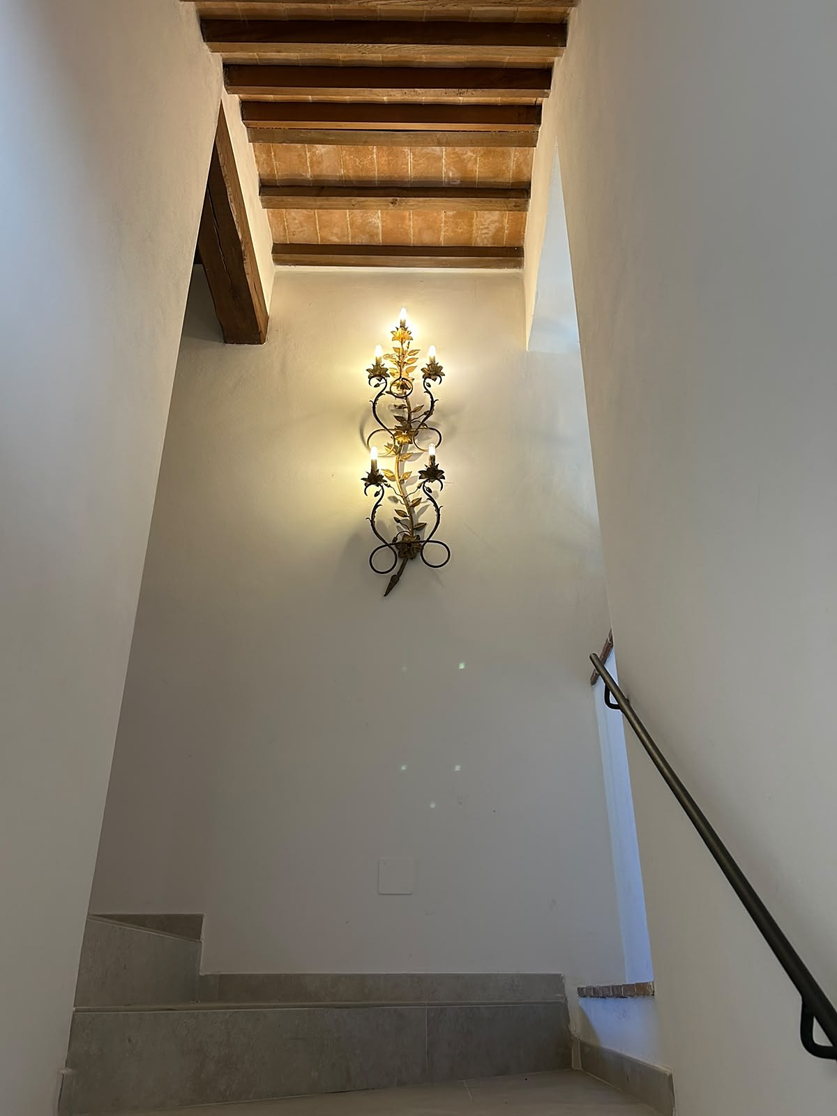 De trap met een vergulde bloemenvormige wandlamp tussen witte muren, onder het houten plafond