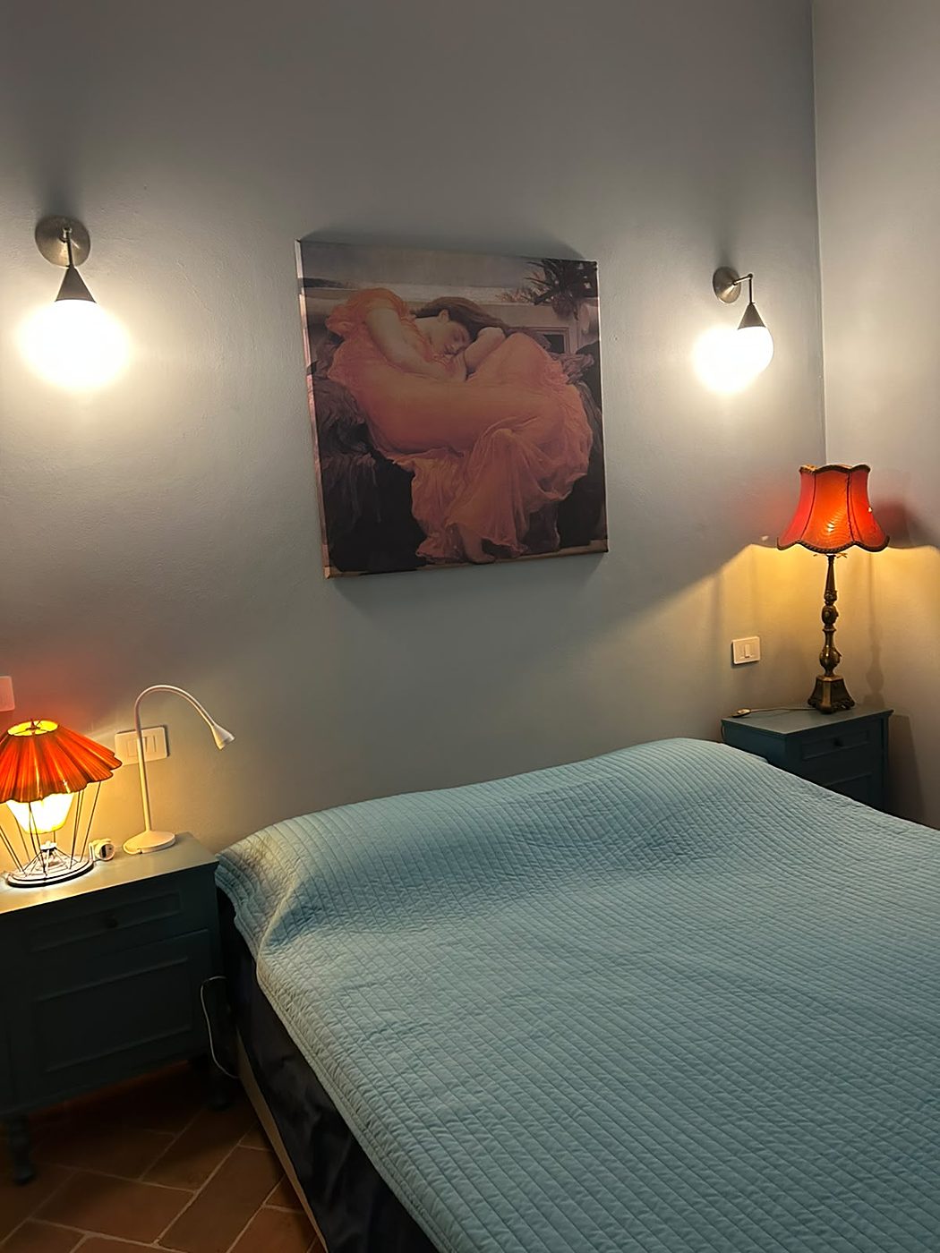 De slaapkamer met een lichtblauw dekbed en oranje nachtlampjes aan de grijze muur
